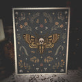 A square art print or card featuring a moth with a skull at its center, surrounded by floral and skull motifs on a dark gray background, styled on a lace-covered surface with dim ambient lighting.