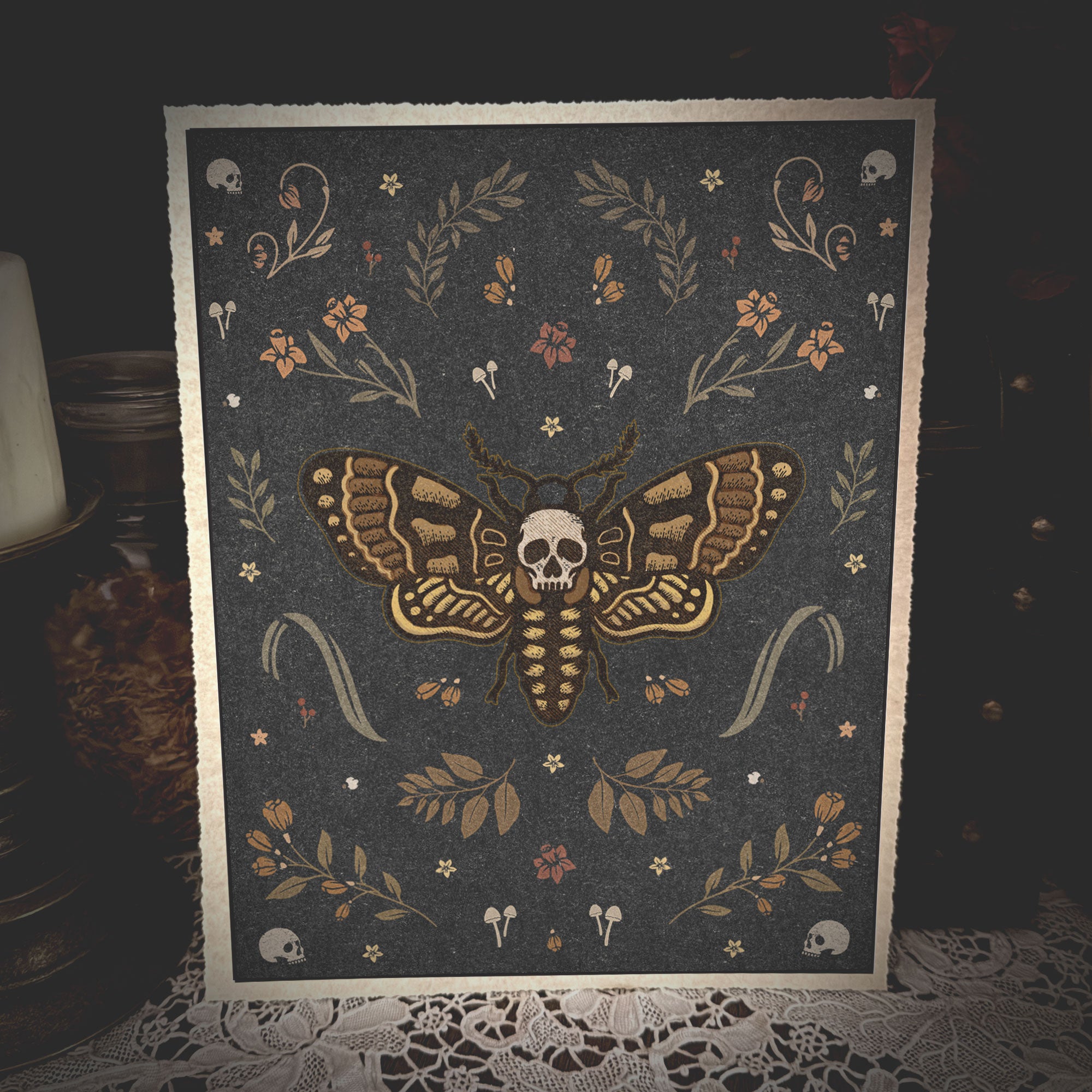A square art print or card featuring a moth with a skull at its center, surrounded by floral and skull motifs on a dark gray background, styled on a lace-covered surface with dim ambient lighting.